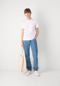 Hvit bomulls t-skjorte med liten rød logo, blå denimjeans, og en lysebeige totebag. Uformelle sko i hvitt med beige detaljer.