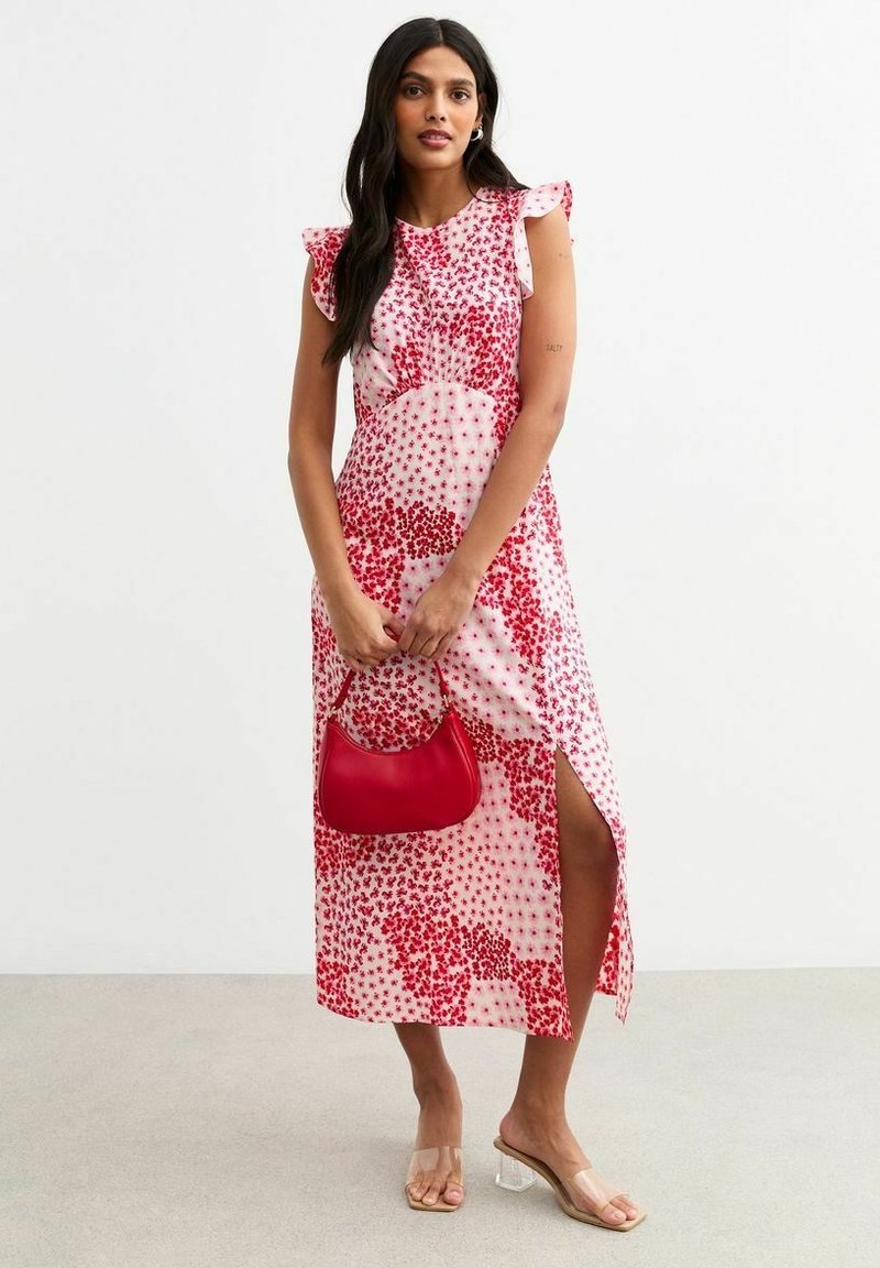 Vestido de estampado floral en blanco con acentos rojos, con mangas rizadas, una abertura lateral y combinado con un bolso rojo y sandalias transparentes.