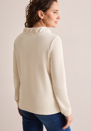 Long-sleeve cream top with a ruffled collar and gathered cuffs, featuring a smooth texture and a relaxed fit, paired with blue jeans.