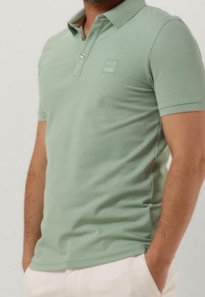 Homme portant un polo à manches courtes vert clair avec un petit logo sur la poitrine, associé à un pantalon beige clair.