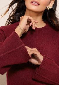 Burgundowy sweter dzianinowy z szerokimi ściągaczami i okrągłym dekoltem. Ręce modelki zdobią srebrna bransoletka i eleganckie kolczyki.