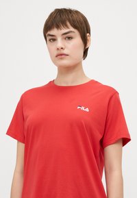 Červené tričko s krátkym rukávom z bavlnenej látky so bielym logom FILA na ľavej hrudi. Hladká textúra a klasický okrúhly výstrih.