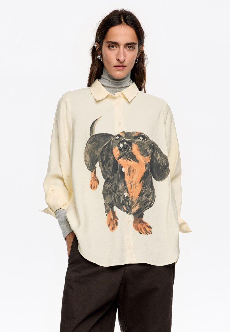 Camisa beige clara con una gran ilustración de un dachshund en negro y naranja, diseño abotonado, con un ajuste holgado y mangas remangadas.