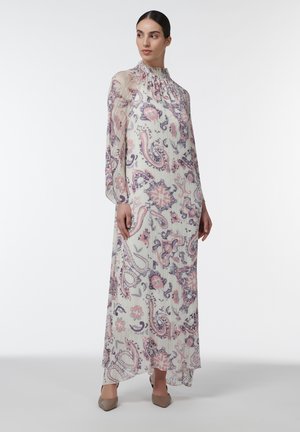 Mujer de pie con un vestido blanco largo y holgado con estampado de paisley en rosa y púrpura, cuello alto, mangas largas y zapatos beige de punta.