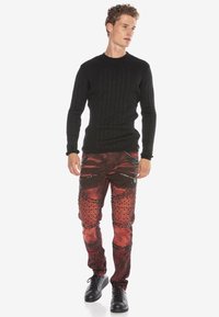 Schwarzer gerippter Strickpullover mit langen Ärmeln, kombiniert mit roten distressed Jeans mit Nietenakzenten und Reißverschlusstaschen. Schwarze Sneakers.