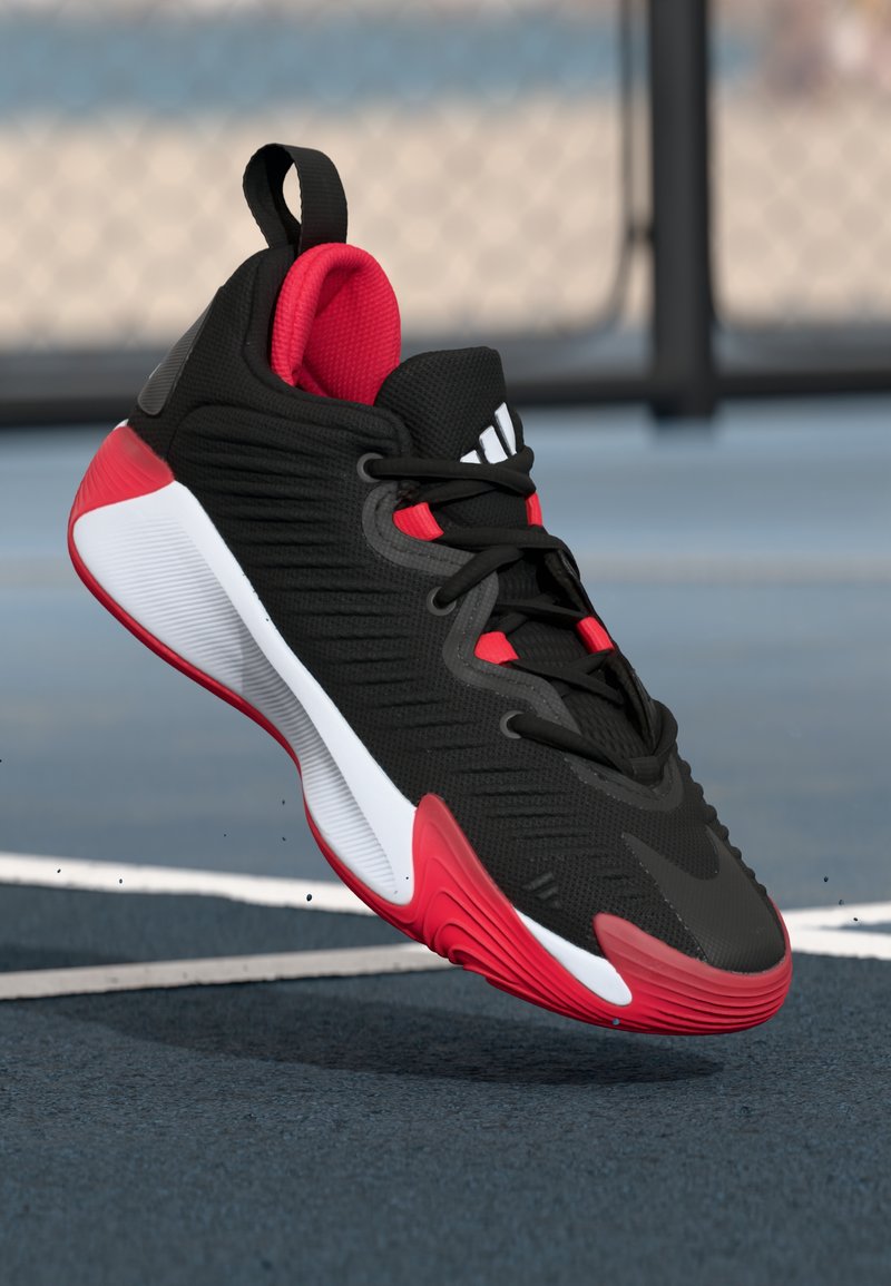 Black and red athletic shoe with white sole, mid-air above blue court surface, with blurred fence in the background.