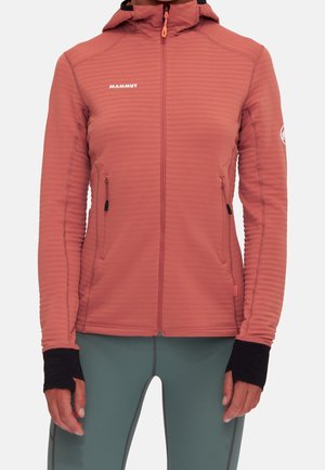 Langarmige koraalkleurige jas met ritssluiting en capuchon, duimgaten in zwarte boorden en met ritssluitingen afgesloten zakken, gedragen door een persoon in een groene broek.
