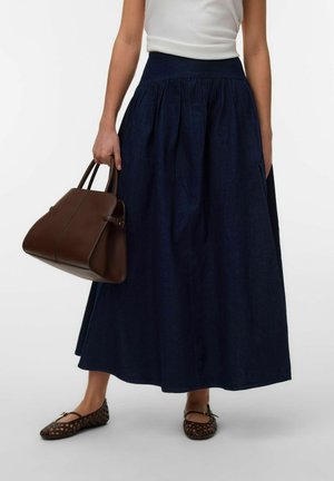 Personne portant un haut blanc sans manches, une longue jupe bleu foncé, des ballerines tressées marron, et tenant un grand sac à main en cuir marron.