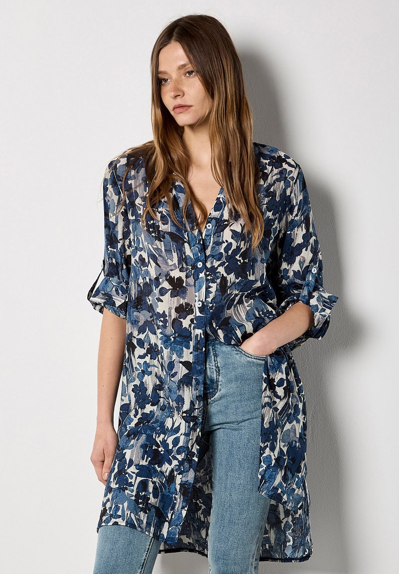 Blouse fleurie bleu et blanc, avec un devant à boutons, manches retroussées et tissu léger et transparent. Coupe ample et décontractée.
