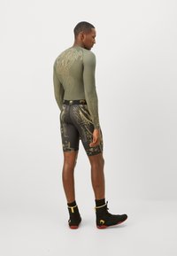 Maglietta sportiva a maniche lunghe color verde oliva con motivo dorato, abbinata a pantaloni corti neri con design astratti. Scarpe alte nere e rosse.