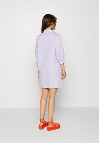 Svetlofialové bavlněné šaty s nabíranými rukávy a límečkem, doplněné oranžovými sandály na řemínky. Hladká tkanina, délka ke kolenům, minimální detaily.