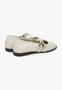 Jasno kremowe płaskie buty ze spiczastym noskiem, z podwójnymi paskami i złotymi detalami. Gładka skórzana tekstura z niskim obcasem.