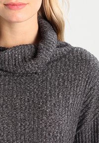 Šedý pletený svetr s vysokým, ohnutým límcem. Má žebrovanou strukturu a mírně volný střih, který poskytuje teplo a pohodlí.