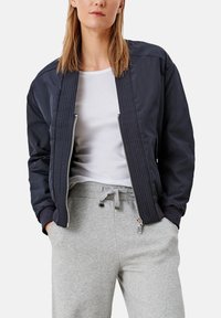 Vrouw in een marineblauwe bomberjack boven een wit t-shirt en grijze joggingbroeken met trekkoord, staande met haar handen in haar zakken.