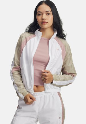 Jonge vrouw in een witte, beige en roze zip-up trainingsjack en bijpassende broek boven een roze crop top, staande tegen een witte achtergrond.