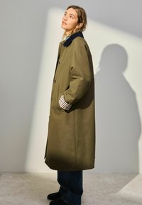 Oliven grøn lang frakke med en marineblå krave, ternede manchetter og sidelommer, med en lige design og glat tekstur.