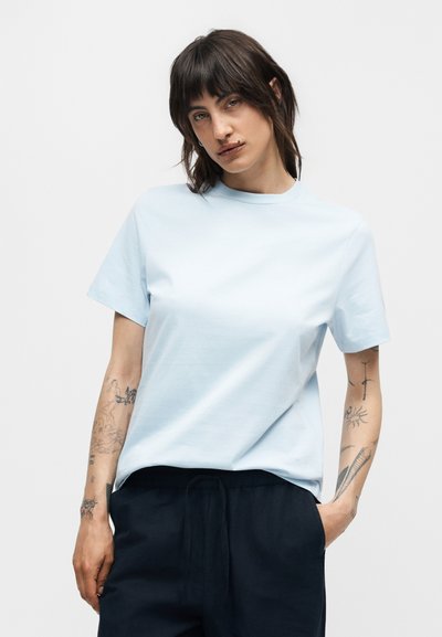 Γυναίκα με σκούρα μαλλιά που φοράει γαλάζιο κοντομάνικο μπλουζάκι και σκούρο παντελόνι, στέκεται με το ένα χέρι στην τσέπη και έχει ορατά τατουάζ στα χέρια.