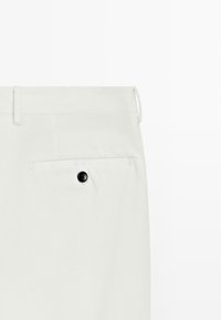Białe spodnie materiałowe o gładkiej fakturze, z tylną kieszenią i czarnym guzikiem. Zaprojektowane w czystym, dopasowanym stylu.