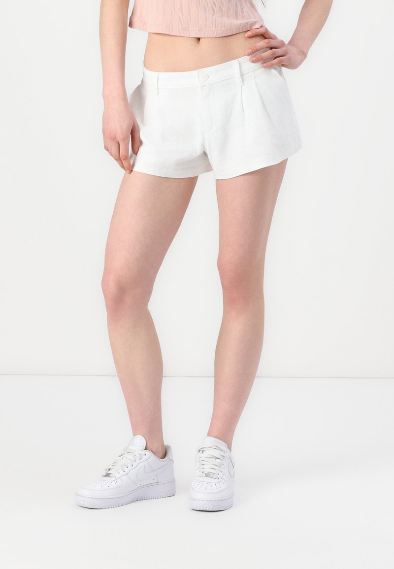 Pantalones cortos de lino blanco con un corte holgado, con pliegues en la parte delantera y cierre de botón, combinados con zapatillas blancas. Textura suave y líneas limpias.