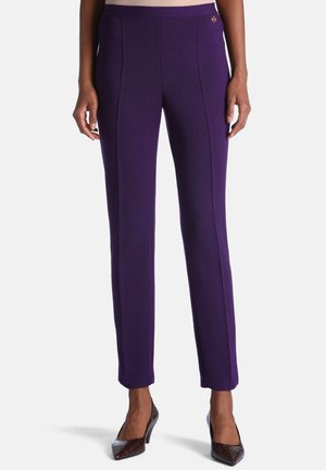Pantalon violet ajusté, coupe droite, porté avec des escarpins à bouts pointus marron foncé sur une personne debout devant un fond uni.