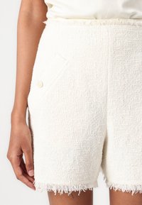 White textured shorts with a frayed hem, featuring a side pocket and a light button accent. Fabric appears soft and woven.