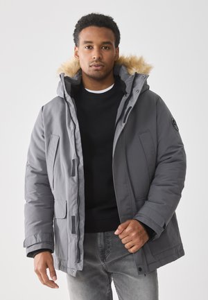 Joven vistiendo una chaqueta de invierno gris con capucha con borde de pelo, sudadera negra y jeans grises, de pie contra un fondo blanco.
