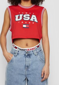 Top corto de malla roja con cuello a rayas en blanco y azul marino, que presenta el logotipo "TOMMY USA JEANS". Jeans de mezclilla azul claro de cintura alta con cinturilla de logo.