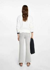 Biely sveter s pletením do vzoru cables a okrúhlym výstrihom, skombinovaný so svetlošedými rozšírenými džínsami. Drží čierny kúsok oblečenia v ľavej ruke.