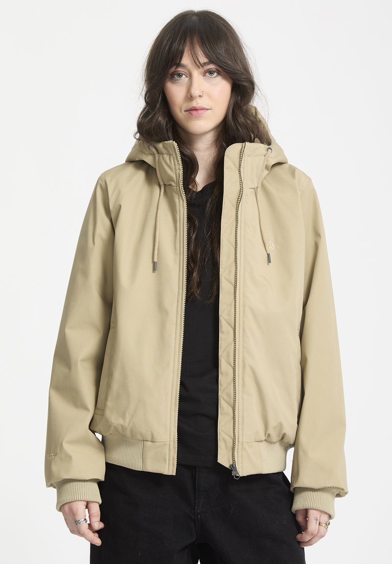 Beige huva jacka med dragkedja, ribbade ärmslut och en slät yta. Har dragsko på huvan och sidofickor.