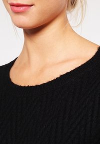 Maglione nero lavorato a maglia con una texture a coste e scollatura ampia. Mostra un materiale morbido e peloso con bordi rifiniti.