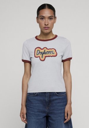 Giovane donna che indossa una t-shirt bianca con rifiniture bordeaux e logo colorato "Drykorn", abbinata a jeans di denim blu, in piedi davanti a uno sfondo semplice.