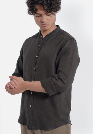 Uomo con capelli ricci che indossa una camicia scura con bottoni e maniche risvoltate, guarda in basso e si sistema il polso sinistro.