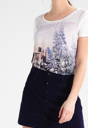 Vrouw draagt een wit, kort mouw topje met een paarse bloemen- en stadsgezichtprint, gecombineerd met een donkere rok met knopen aan de voorkant.