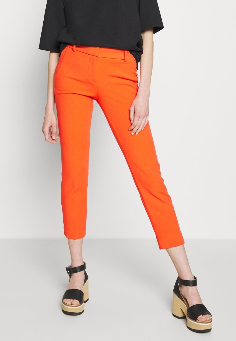 Pantalon court orange à coupe ajustée, avec une texture lisse. Associé à des sandales à talons carrés noires et un haut noir ample.