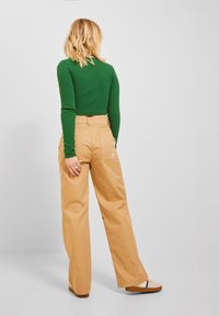Zielona bluzka z długim rękawem w połączeniu z beżowymi spodniami o szerokich nogawkach. Spodnie mają tylną kieszeń oraz subtelną logo blisko pasa.