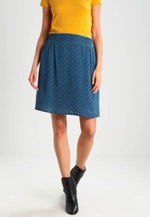 Woman wearing a blue patterned knee-length skirt, mustard yellow short-sleeve top, and black ankle boots standing on a white background.