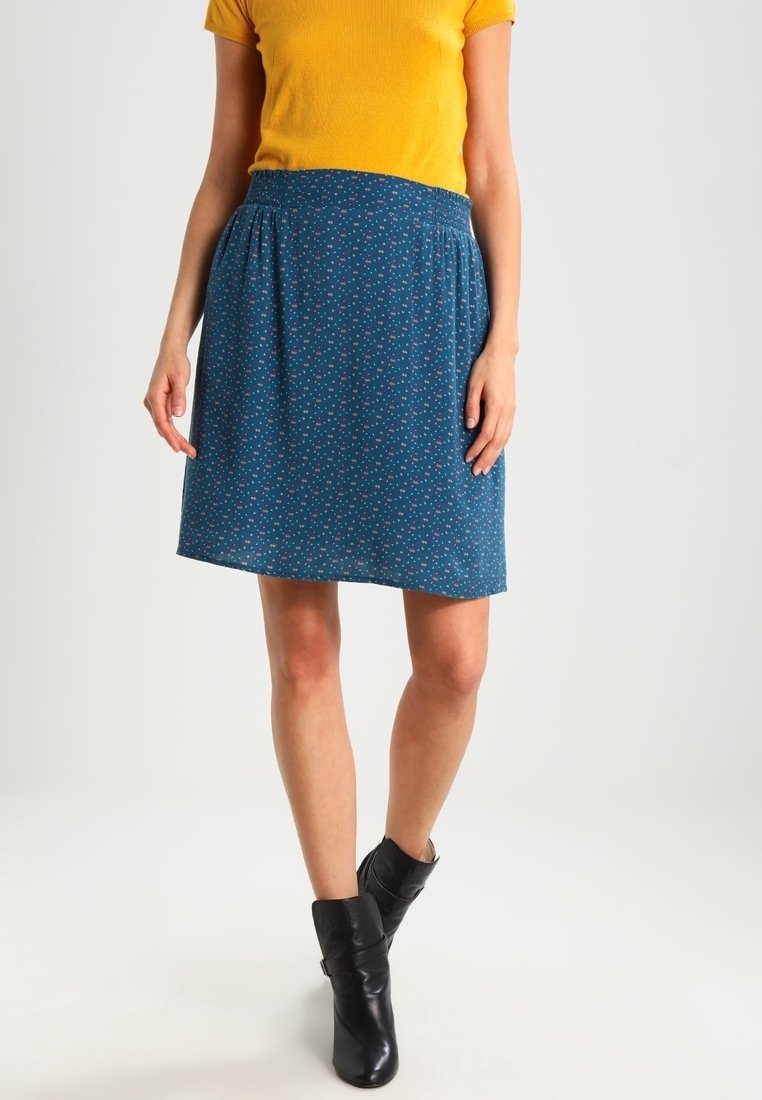 Femme portant une jupe bleue à motifs longueur genoux, un haut à manches courtes jaune moutarde et des bottines noires, debout sur un fond blanc.