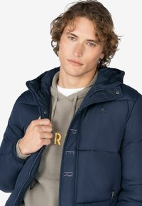 Marine blauwe pufferjack met een afneembare capuchon, ritssluiting aan de voorkant en elastische manchetten. Draagt over een beige hoodie met een gestructureerd patroon en gouden letters.