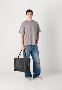 Pilkas trumpas moteriškas marškinėliai su "Calvin Klein" logotipu, derinami su nusidėvėjusiais mėlynais džinsais ir smėlio spalvos sportbačiais. Modelis laiko juodą odinį rankinį krepšį.