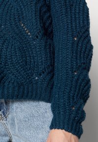 Teal stickad tröja med ribbad mudd och ett strukturerat mönster med öppna detaljer. Kombinerad med ljusblå denim.