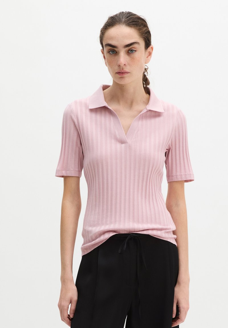 Frau mit geflochtenem Haar, die ein hellrosa geripptes Kurzarm-Poloshirt und schwarze Kordelhose trägt, steht vor einem weißen Hintergrund.