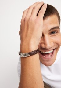 Pulsera de cuero trenzado marrón con cierre de metal plateado, mostrando un tejido intrincado y un acabado liso y pulido.