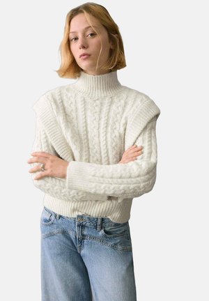 Suéter blanco de punto con cuello alto y patrón de trenzas, combinado con jeans de mezclilla azul claro. Diseño texturizado y voluminoso con puños de canalé.