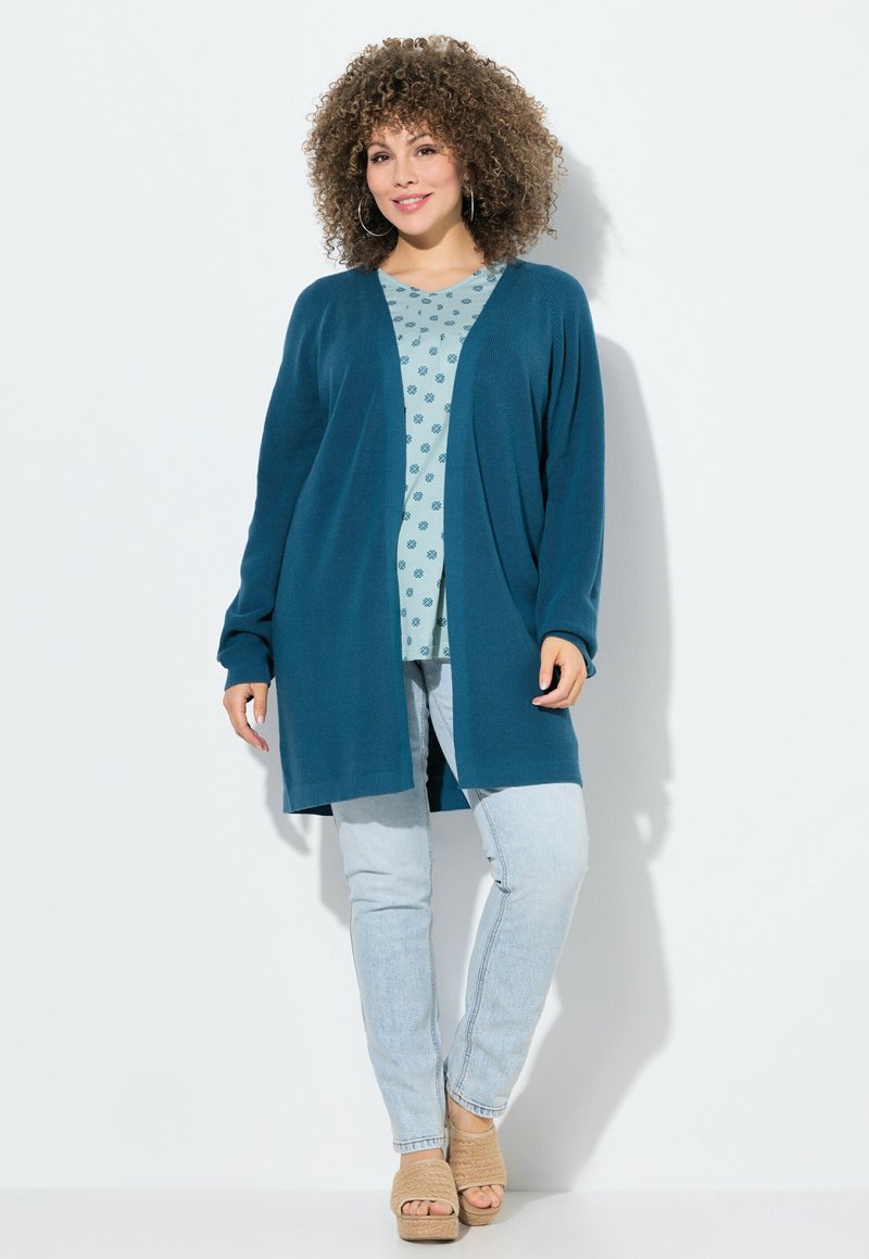 Cardigan largo en color verde azulado con frente abierto, textura de punto, llevado sobre una blusa de estampado azul claro, combinado con jeans claros y sandalias de cuña.