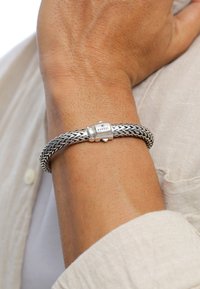 Bracciale intrecciato in argento con chiusura cilindrica incisa con la scritta "Kuzjoi". Indossato su un polso abbronzato, evidenziando una texture intricata.