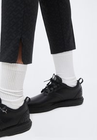 Πόδια που φορούν μαύρα χαμηλά αθλητικά παπούτσια και λευκές κάλτσες, με μαύρο παντελόνι cropped που έχει φερμουάρ στο πλάι.