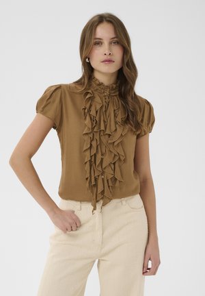 Frau mit welligem Haar, die eine braune Bluse mit kurzen Rüschenärmeln und cremefarbene High-Waist-Hosen trägt und mit einer Hand in der Tasche steht.