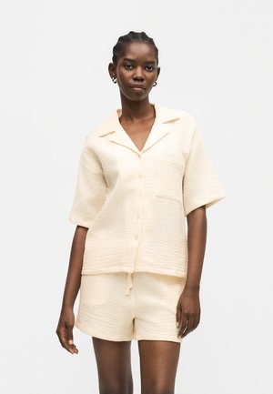 Vrouw draagt cremegekleurde, gestructureerde korte mouwen overhemd met kraag en knopen en bijpassende korte broek tegen een effen achtergrond.
