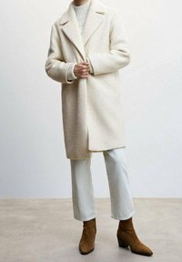 Persona vistiendo un abrigo texturizado crema, un suéter crema de canalé, pantalones de color claro y botines de ante marrón, de pie sobre un suelo neutro.