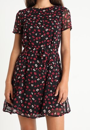 Femme portant une robe noire à manches courtes avec un motif floral rouge et blanc ainsi qu'une ceinture à la taille, debout devant un fond uni.